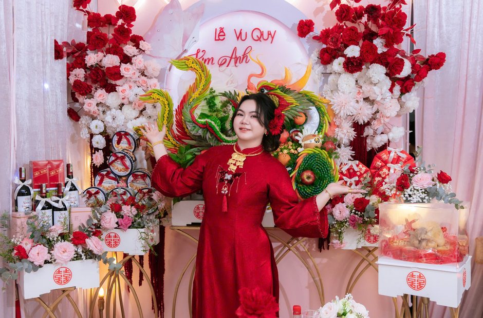 Cô dâu mặc áo dài đỏ trong lễ vu quy bên mâm quả và phông hoa đỏ
