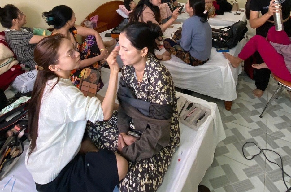Nhiều người cùng được trang điểm trong một căn phòng, thợ đang đánh nền cho một người