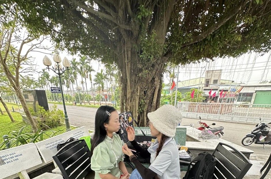 Hai người ngồi trang điểm ngoài trời dưới gốc cây lớn cạnh vỉa hè