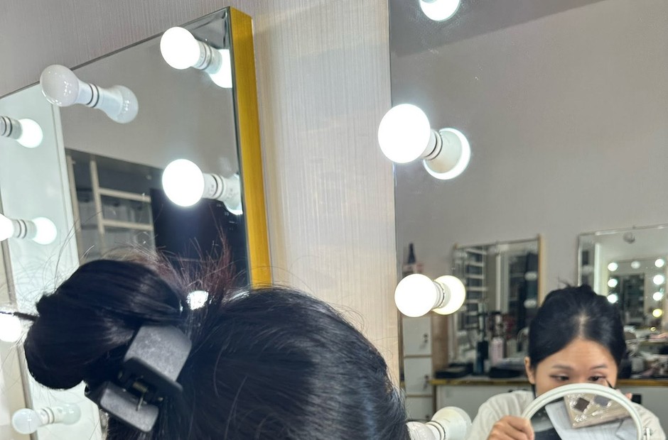 Người ngồi trước gương có đèn viền, cầm gương nhỏ trang điểm mắt, tóc búi kẹp phía trước